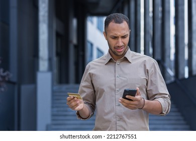 Frustrated And Upset African American Man Outside Office Building Trying To Make A Purchase In An Online Store, Man Using Smartphone And Bank Credit Card, To Transfer Money And Buy Online