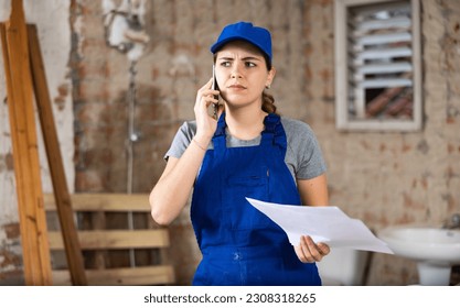 Frustrated woman builder in overall standing in construction site and talking on phone about project documentation. - Powered by Shutterstock