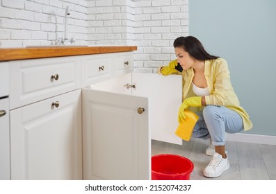 Frustrated woman on phone calls plumber because of leaking pipe in sink in kitchen. Young housewife in yellow rubber gloves sits near sink with bucket and washcloth and calls plumbing repair service.