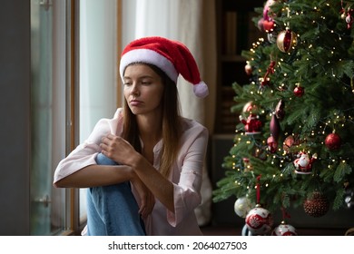 Frustrated Xmas young woman getting bored at Christmas tree, sitting at window at home, thinking over problems, feeling lonely, sad, depressed, suffering from apathy, loneliness on holiday time