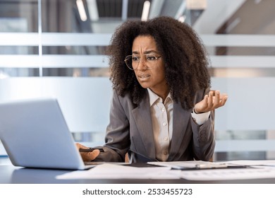 Frustrated young african american business woman sitting in office at work desk with documents and looking at laptop screen in frustration with arms outstretched. - Powered by Shutterstock