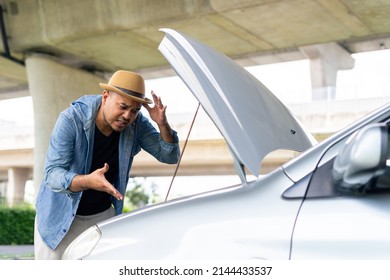 Frustrated young asian man looking at broken down car engine on street waiting for assistance to arrive and recover or fix the vehicle. Male angry stand front a broken car. Broken car while traveling.