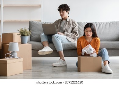 Frustrated Young Asian Woman Unpacking Carton Box Alone While Her Lazy Boyfriend Sitting On Couch With Laptop Pc, Copy Space. Moving Day, Relocation And Family Conflicts Concept