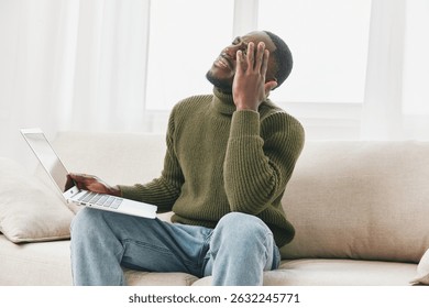 A frustrated young Black man in a green turtleneck sitting on a couch, holding his laptop in one hand and rubbing his forehead, depicting a sense of anxiety and stress