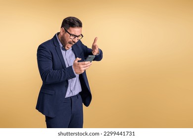 Frustrated young businessman looking and pointing at smart phone while screaming angrily. Male professional yelling aggressively while reading message isolated on beige background - Powered by Shutterstock