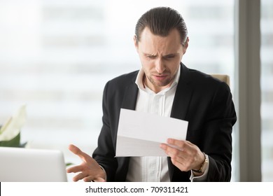 Frustrated Young Businessman In Suit Looking At Confusing Letter In Modern Office Setting. Unexpected High Bill, Unpaid Debt, Failing Financial Report, Tax Delinquency, Breach Of Contract Concept.