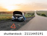 A frustrated young girl stands near a broken-down car in the middle of the highway during sunset. Breakdown and repair of the car. Waiting for help. Car service. Car breakdown on road.