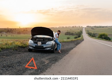 A frustrated young girl stands near a broken-down car in the middle of the highway during sunset. Breakdown and repair of the car. Waiting for help. Car service. Car breakdown on road.