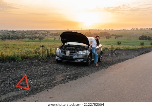 A
frustrated young girl stands near a broken-down car in the middle
of the highway during sunset. Breakdown and repair of the car.
Waiting for help. Car service. Car breakdown on
road.