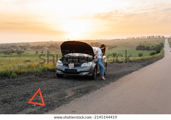 A\
frustrated young girl stands near a broken-down car in the middle\
of the highway during sunset. Breakdown and repair of the car.\
Waiting for help. Car service. Car breakdown on\
road.
