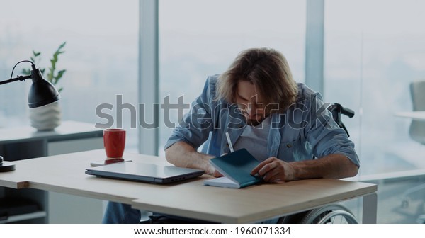 Frustrated young hesitating man using\
computer laptop sitting by desk doubting what to do. Self-doubting\
disabled man in wheelchair working first day at\
office.