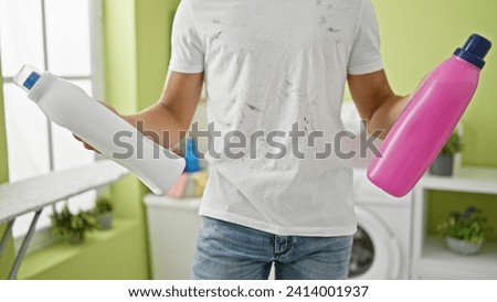 Frustrated young hispanic teenager, sporting a dirty t-shirt, holding detergent bottles whilst tackling laundry chores in a dingy home laundry room.