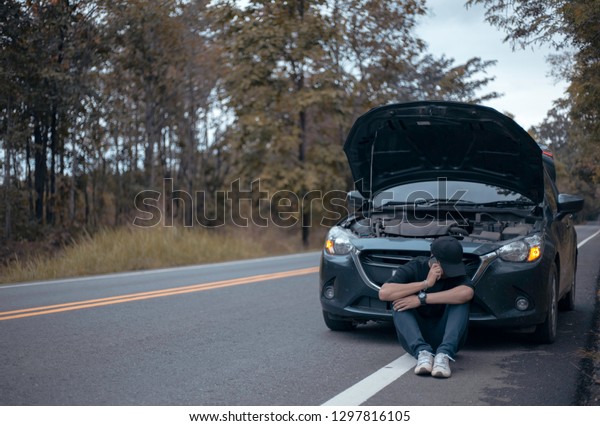 frustrated young man driver phoning for\
assistance while sitting on the road near to broken car. Road trip\
problems and assistance\
concepts.