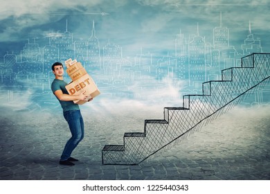 Frustrated Young Man Too Much Burden, Lean Back Carrying Heavy Boxes With Debt Word Text, Have To Climb A Staircase. Concept Of Difficult Life Road, A Lot Of Credits And Taxes To Pay.