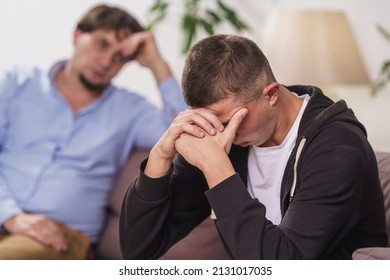 frustrated, young man is sitting at home on couch. difficult teenager covers his face with his hands, he has problems. father talks to his son, discusses problems, supports him. Transitional age - Shutterstock ID 2131017035