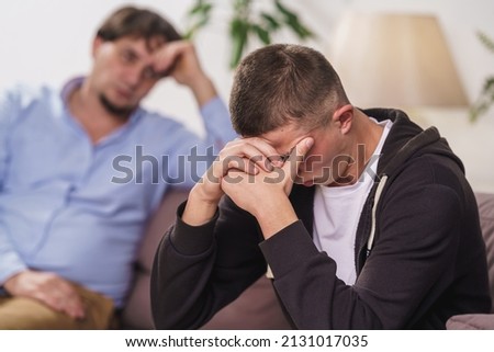 frustrated, young man is sitting at home on couch. difficult teenager covers his face with his hands, he has problems. father talks to his son, discusses problems, supports him. Transitional age