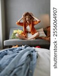 A frustrated young woman with curly hair sitting on a couch, surrounded by messy clothes, embodying the stress of tidying up and organization