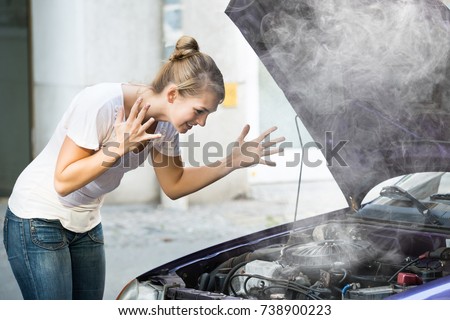 Frustrated Young Woman Looking Under The Hood Of Breakdown Car