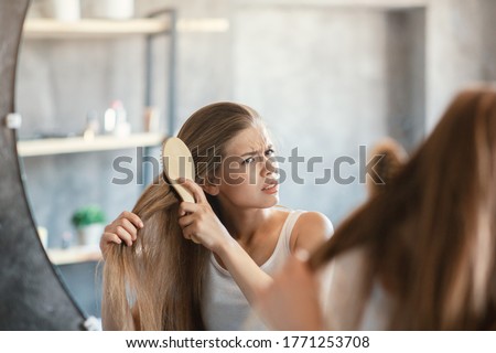 Frustrated young woman trying to brush her tangled hair in front of mirror at bathroom