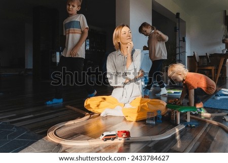 Frustrating woman alone struggling burnout with kids. Stressed out mother sitting on floor in middle of toys while children naughty running around her at room. Family home with chaos, mess.