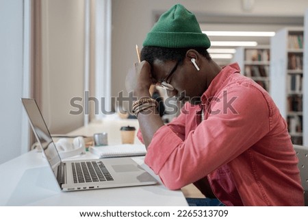 Frustration in distance education. Frustrated young African American guy sitting in front of laptop in library, struggling with online learning, tired male student feeling unmotivated to study