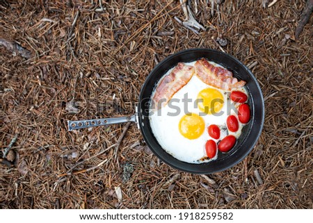 Frying pan with fried eggs, bacon and tomatoes in nature. Cooking breakfast on a gas stove at a summer camp.