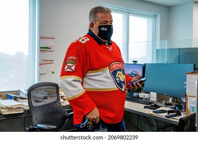 Ft. Lauderdale, Florida, USA - October 29th, 2021 - Overweight White Man Wearing Florida Panthers Jersey Behind Computer Cubicle Farm Desk And Office Chair Standing Wearing Face Mask Holding Cellphone