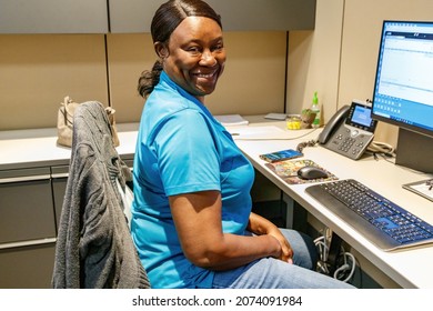 Ft. Lauderdale, Florida, USA - October 29th, 2021 - African American Middle Aged Employee Woman At A Cubicle Work Desk With Phone Sitting In Office Chair Working On Computer Looking At Camera Smiling.