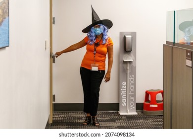 Ft. Lauderdale, Florida, USA - October 29th, 2021 - Female Woman Employee Holding Door Near Hand Sanitizing Dispenser In A Cubicle Farm Wearing Witch Hat, Face Mask, Orange Shirt Costume For Halloween