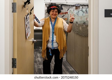 Ft. Lauderdale, Florida, USA - October 29th, 2021 -Female Employee Woman Wearing A Hippie Costume For Halloween With Wig Holding Door Near Cubicle Farm At Work During Office Party With Decorations 