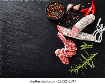 Fuet Sausage Cut In Slices On A Black Slate Plate With Peppercorns, Sliced Chili And Fresh Rosemary, View From Above, Close-up