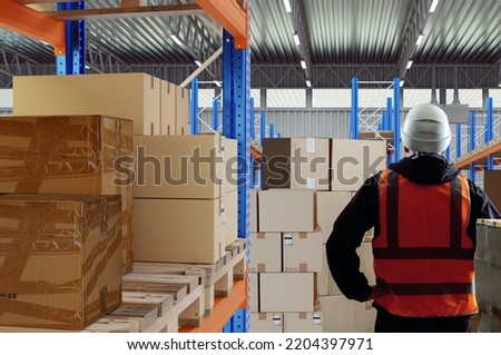 Fulfillment center. Man works for fulfillment company. Shelves with boxes behind man. Warehouse fulfillment employee. Parcels with stickers on pallets. Guy in white helmet with his back to camera