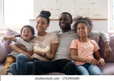 Full African ethnicity family, spouses with daughter and son spend weekend free time together at home sitting on couch in living room eating popcorn watching TV show movie or cartoons have fun concept