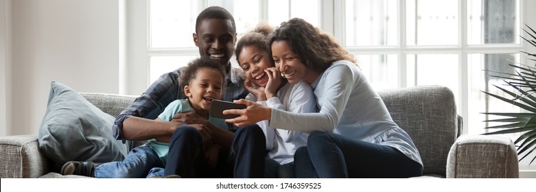 Full African family with little kids sit on couch in living room having fun using smart phone new cool application, taking selfie, watch funny videos. Horizontal photo banner for website header design