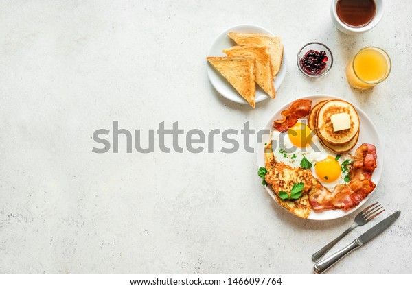 Full American Breakfast on white,\
top view, copy space. Sunny side fried eggs, roasted bacon, hash\
brown, pancakes, toasts, orange juice and coffee for\
breakfast.