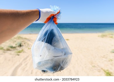 A Full Bag Of Trash In A Man's Hand Against The Backdrop Of A Clean Beach And Sea. Garbage Collection Concept. Selective Focusing.