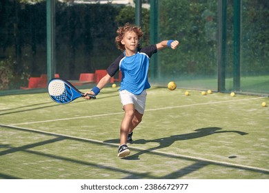 Full body of active boy in activewear hitting ball with racket while playing padel game on sports ground on summer day