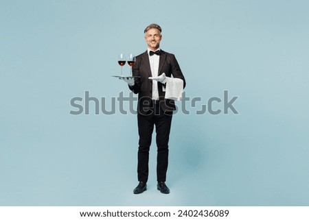 Full body adult barista sommelier male waiter butler man wear shirt black suit bow tie uniform hold tray two glasses of wine work at cafe isolated on plain blue background. Restaurant employee concept