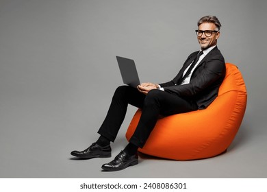 Full body adult smiling employee business man corporate lawyer wear classic formal black suit shirt tie work in office sit in bag chair hold use laptop pc computer isolated on plain grey background - Powered by Shutterstock