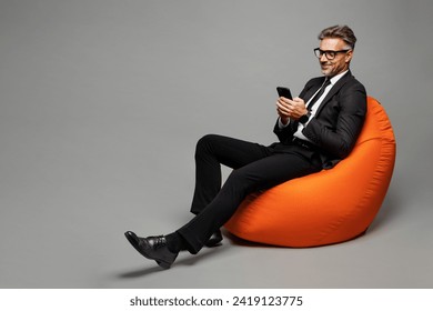 Full body adult successful employee business man corporate lawyer wear classic formal black suit shirt tie work in office sit in bag chair hold use mobile cell phone isolated on plain grey background - Powered by Shutterstock