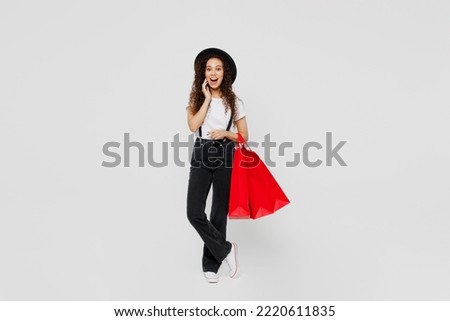 Full body amazed shocked happy fun young woman wear t-shirt hat hold in hand red paper package bags after shopping touch face isolated on plain solid white background Black Friday sale buy day concept