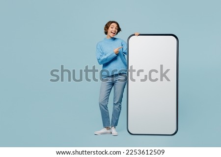 Full body amazed young woman wears knitted sweater point index finger big huge blank screen mobile cell phone smartphone with workspace mockup area isolated on plain pastel light blue cyan background