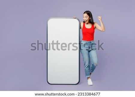Full body amazed young woman of Asian ethnicity wear casual clothes red tank shirt big huge blank screen mobile cell phone with area do winner gesture isolated on plain pastel light purple background