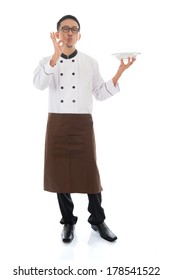 Full Body Asian Chef Holding An Empty Plate, Showing Tasty And Satisfied Hand Sign, Standing Isolated On White Background.