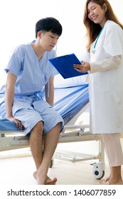 Full Body Asian Female Doctor Smiling Explains The Symptoms To A Young Male Patient. A Sick Man Sitting On A Hospital Bed.