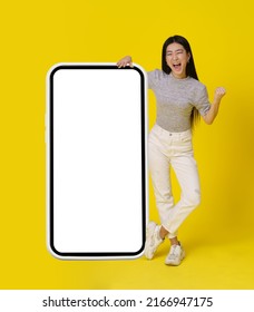 Full Body Asian Girl Stand Near Huge, Giant Smartphone With White Screen Happy Making Yeah, Winner Hand Gesture Dressed In Casual Isolated On Yellow Background. Free Space Mock Up. 