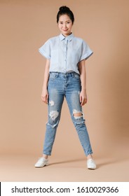 Full Body Of An Asian Woman In Light Blue Short Sleeve Shirt And Blue Jeans,  Little Smiling Face Stand On Studio Beige Background.