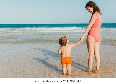 Full Body Back View Of Cheerful Young Mother In Swimwear Holding Hand Of Little Shirtless Toddler Kid While Standing Together On Sandy Beach Near Waving Sea In Summer 