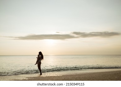 Full Body Back View Of Cheerful Young Mother In Swimwear Holding Hand Of Little Shirtless Toddler Kid While Standing Together On Sandy Beach Near Waving Sea In Summer, Sunset Time