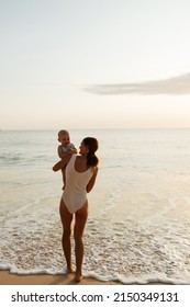 Full Body Back View Of Cheerful Young Mother In Swimwear Holding Hand Of Little Shirtless Toddler Kid While Standing Together On Sandy Beach Near Waving Sea In Summer, Sunset Time